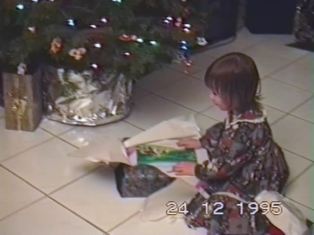 A vintage VHS recording from Christmas Eve 1995 shows a little girl in a festive dress opening a present next to the Christmas tree