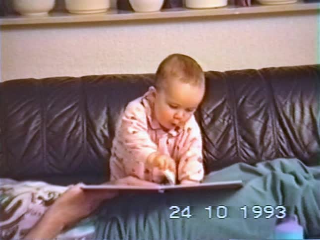 A cute baby in pink pajamas sits on a couch next to their grandfather and curiously flips through the pages of a picture book in this vintage home video from 1993