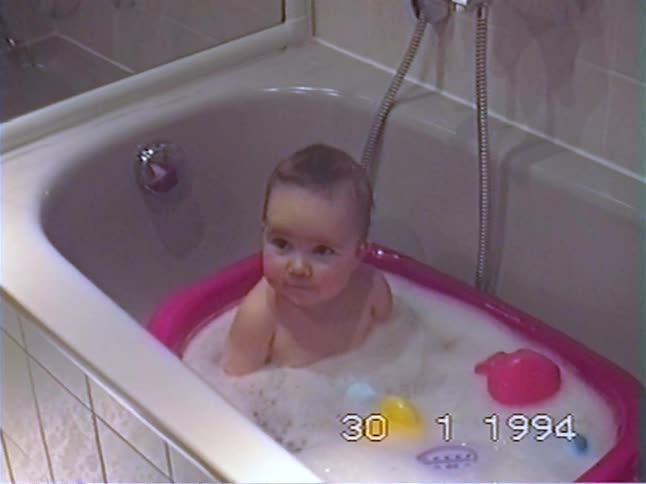 An adorable baby sits in a pink inflatable tub, playing in a bubble bath before looking up with a big smile