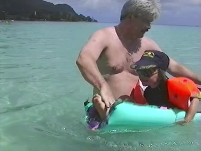Man helps a child with orange floaties on an inflatable raft in clear tropical ocean water