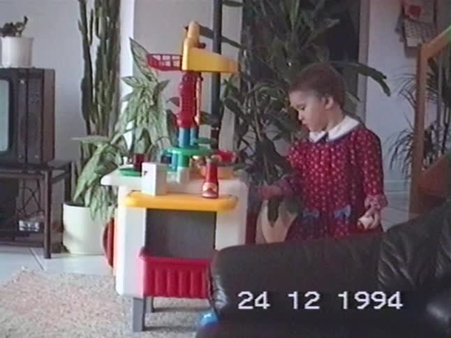 A little girl in a red dress plays with her new toy kitchen on Christmas Eve in a candid 1994 home video