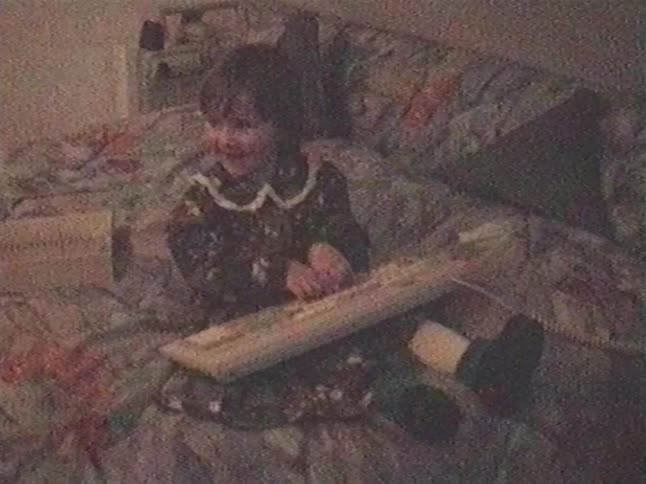 A cute little girl sits on her bed and happily types on an old computer keyboard in a grainy, nostalgic home video