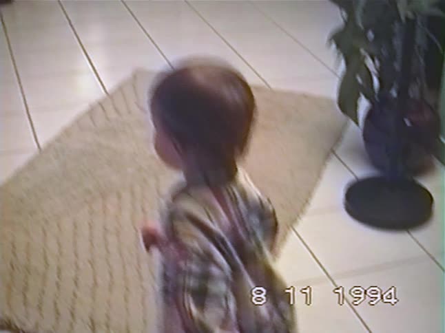 A cute toddler wearing a plaid shirt energetically dances in the living room in this vintage home video from 1994
