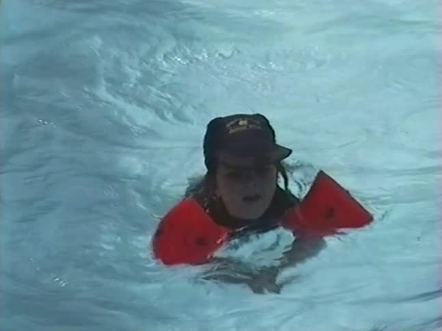 A vintage home video shows a happy young girl with red water wings floating in a swimming pool