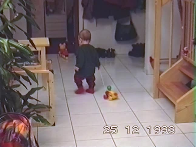 A sweet toddler in a Christmas sweater walks down a hallway pulling a toy on Christmas morning in 1993