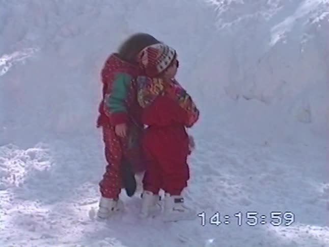 Two small children in colorful 1990s snowsuits hug each other while playing in the deep snow