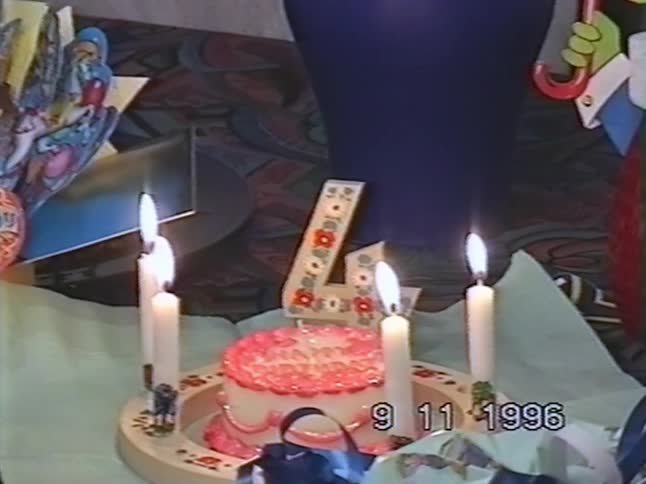 A nostalgic close-up from 1996 reveals a 4th birthday cake with four lit candles, slowly pulling back to show wrapped presents and decorations