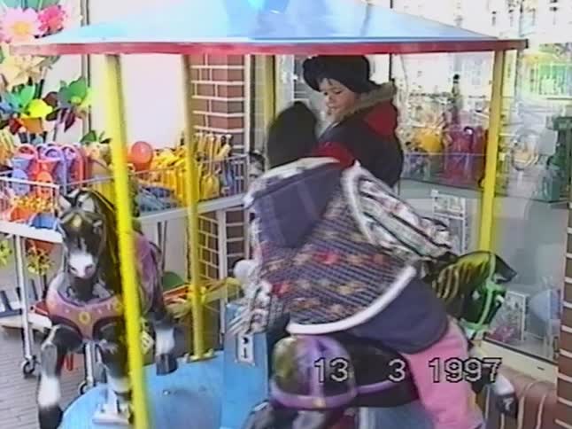 Vintage 1997 VHS footage shows two happy children in winter coats riding a colorful carousel outside a toy store