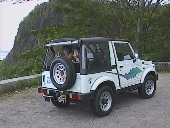Vintage home movie captures friends waving from the back of a convertible SUV on a scenic mountain road trip