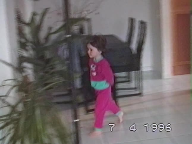 A young girl in pink pajamas playfully peeks out from the kitchen and hides behind a plant in a 1996 home video