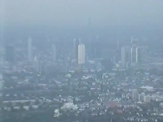 Vintage 1980s aerial footage captures a hazy, slow zoom towards the Frankfurt city skyline on an overcast day