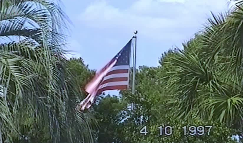 Vintage home movie footage from 1997 shows an American flag waving gently in the breeze, framed by lush palm trees against a blue sky