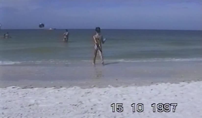 A young girl in a swimsuit walks to the edge of the calm ocean on a white sand beach in this nostalgic VHS clip from 1997