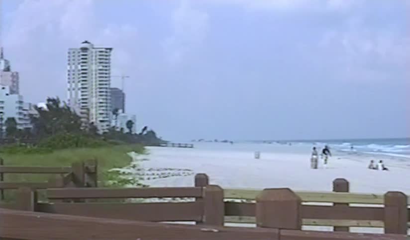 A vintage VHS style panning shot reveals the iconic Art Deco hotels and sandy shores of Miami Beach on a cloudy day