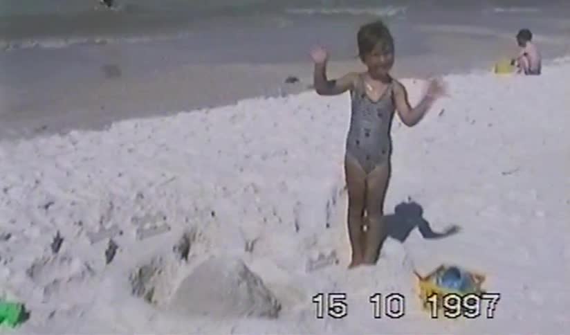 A happy young girl waves at the camera next to her sandcastle on the beach in this nostalgic 1997 home video