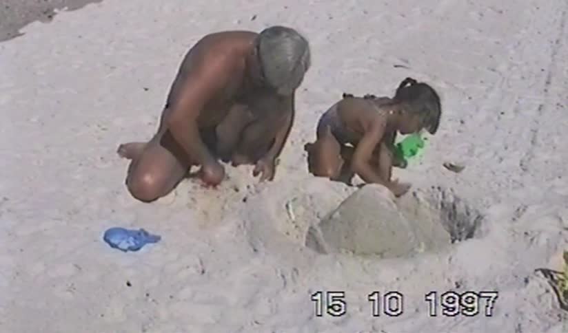 A grandfather and his granddaughter happily build a sandcastle together on a sunny beach in this vintage home movie from 1997