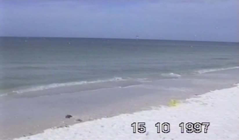 A nostalgic home video from 1997 captures a little girl on a white sand beach before panning to the ocean and the crowded shoreline