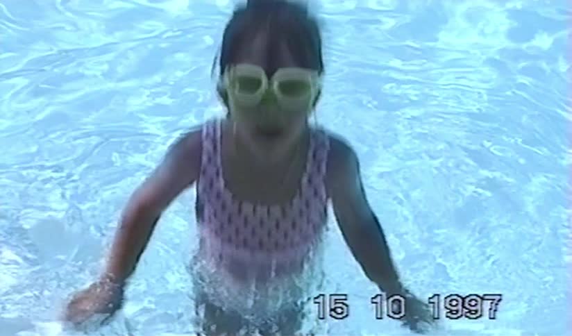A young girl with green goggles learns to swim in a pool in a vintage home video from 1997