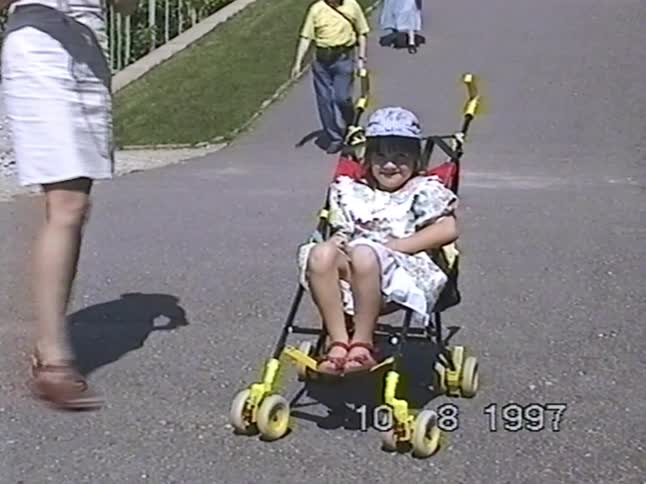 A vintage 1997 home video captures a happy little girl in a stroller smiling at the camera while her mother walks up beside her
