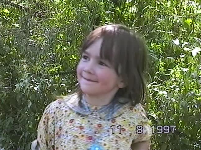 A vintage 1997 home movie captures a cute little girl in a floral dress smiling shyly for the camera in a garden
