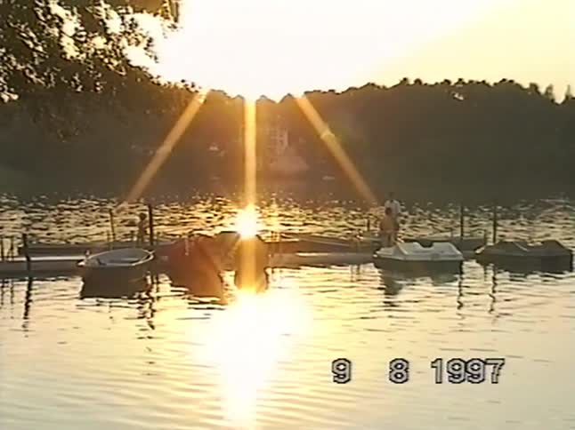 A nostalgic 1997 VHS clip shows a serene sunset reflecting off a calm lake with people standing on a dock next to paddle boats