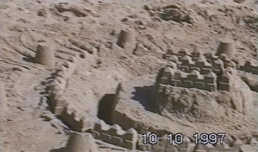 A vintage home video from 1997 shows a young child finishing an elaborate sandcastle on a sunny beach