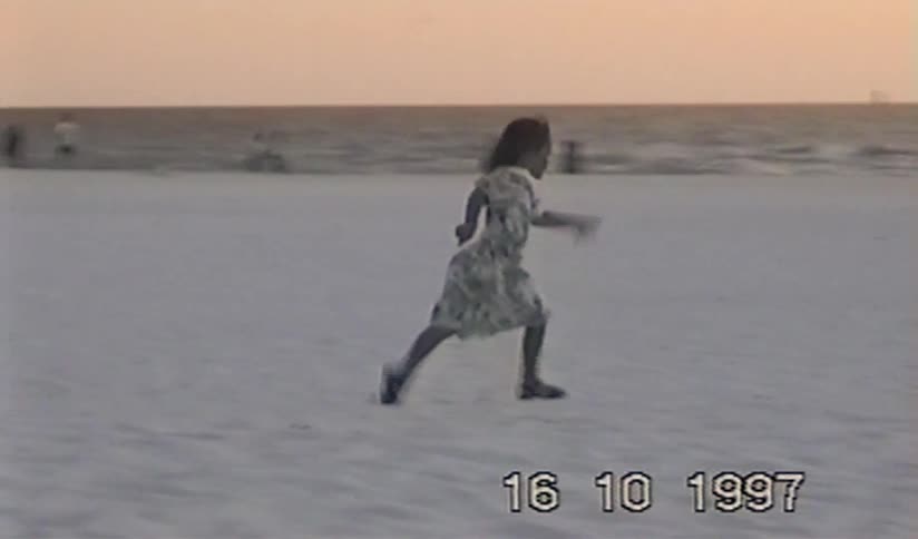 A young girl in a pretty dress playfully runs and dances on a sandy beach at sunset in this vintage 1997 home movie