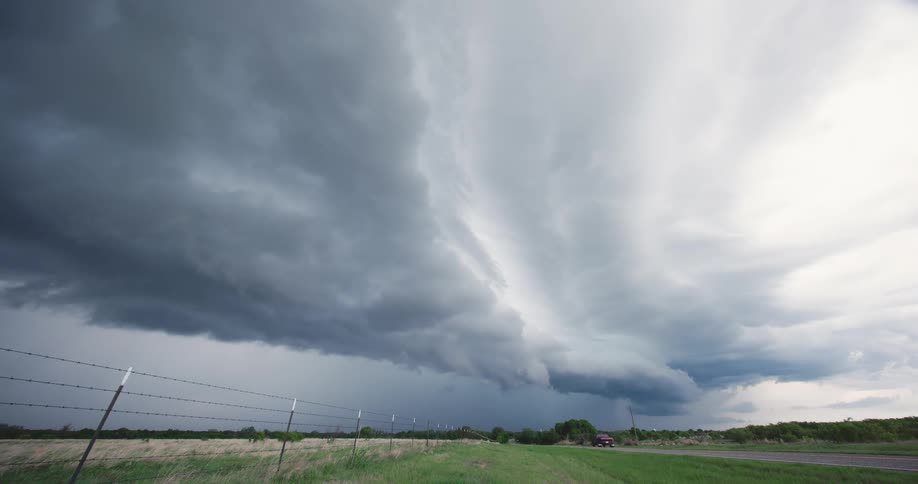 Dark, dramatic supercell storm clouds with occasional lightning flashes hover over a vast rural landscape with a country road
