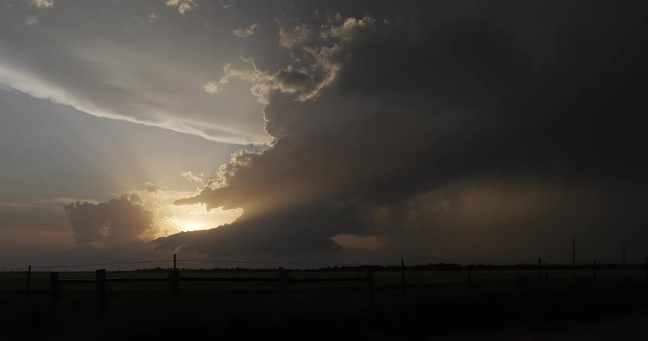 Sun rays beam through dramatic storm clouds gathering at sunset over a silhouetted rural landscape