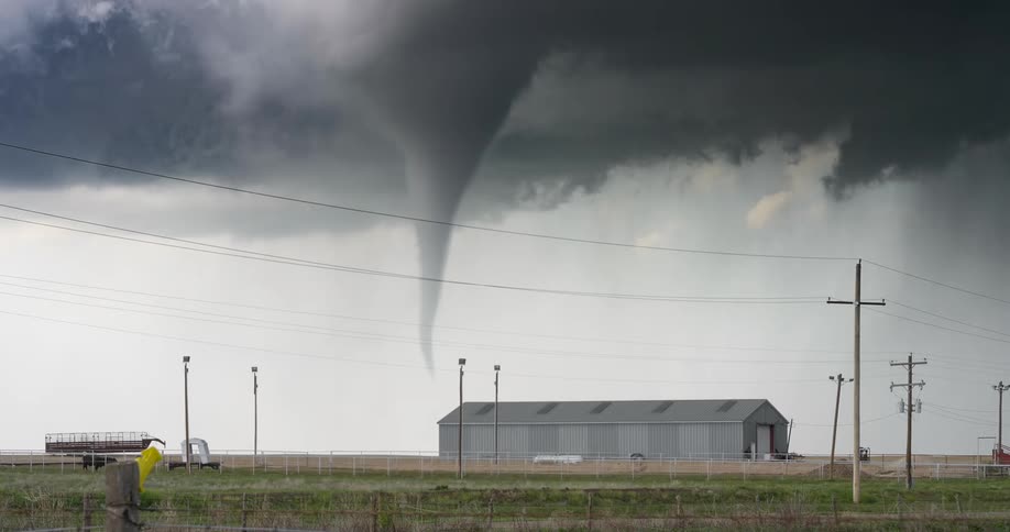 A powerful tornado descends from a dark supercell storm cloud over a rural farm building as a car drives by