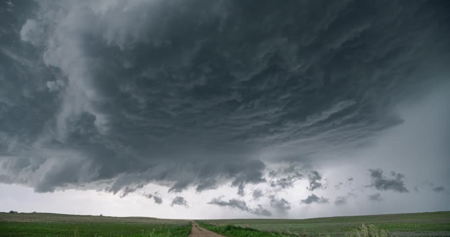 A massive, swirling supercell storm cloud looms ominously over a green rural landscape and a dirt road