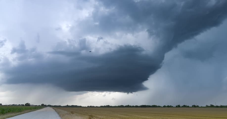 A massive, ominous supercell thunderstorm looms over a rural landscape with a dirt road and farm fields