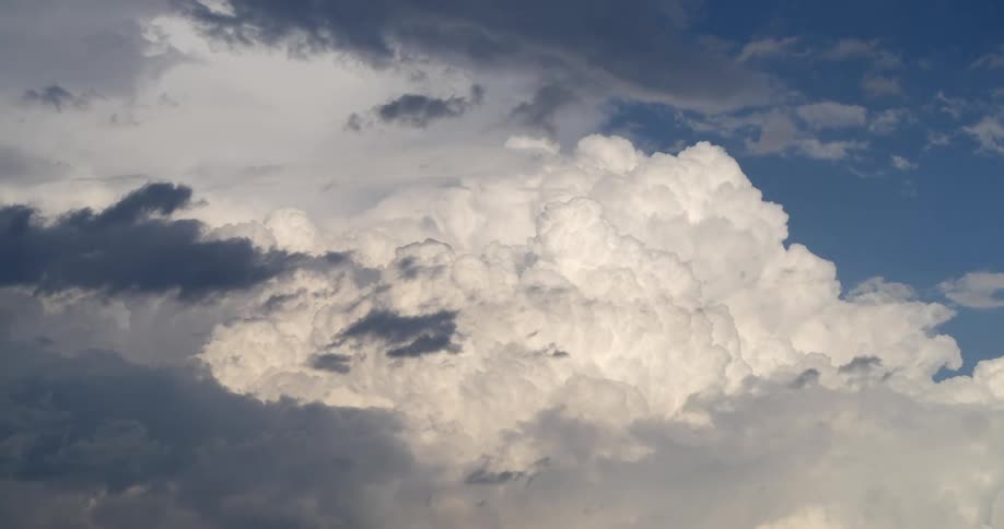 A majestic and powerful white cumulonimbus cloud billows slowly in a blue sky, framed by dark, ominous storm clouds