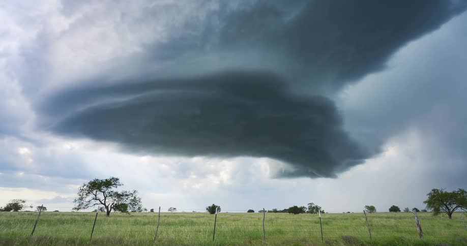 A massive supercell thunderstorm with a rotating updraft looms ominously over a green rural field