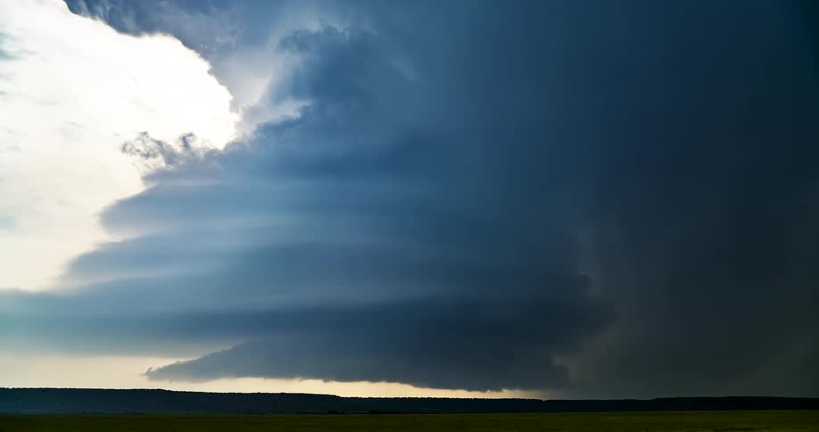 A massive supercell thunderstorm with a dramatic, layered structure looms over a flat, green landscape as lightning flashes inside