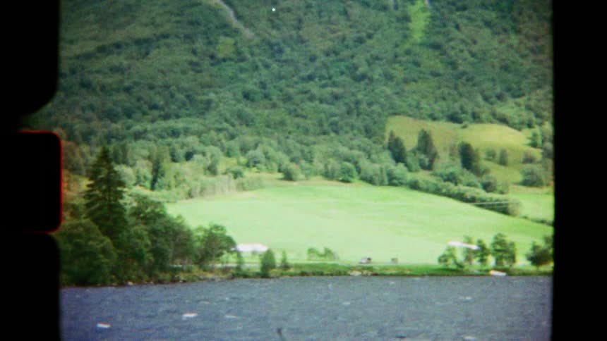 Vintage 8mm film captures the serene view of a vast lake bordered by lush green mountains and quaint houses