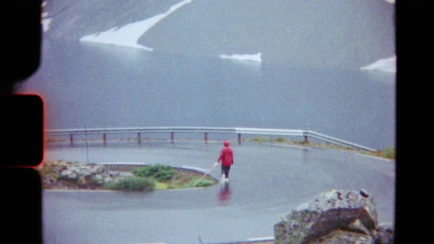 Vintage 8mm footage of a person in a red raincoat walking along a winding mountain road overlooking a snowy fjord on a foggy day