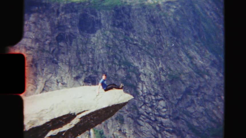 Vintage 8mm footage shows a lone hiker sitting on the edge of the iconic Trolltunga rock formation, overlooking a majestic Norwegian fjord