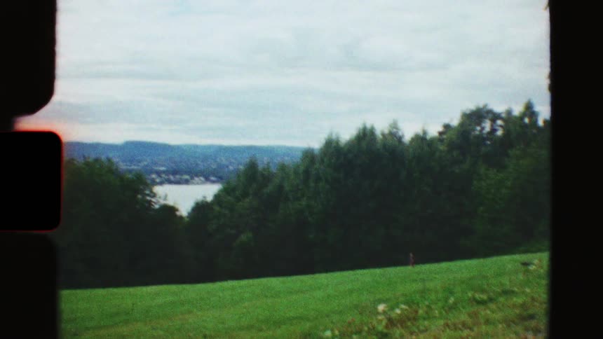 Nostalgic 8mm film captures a lone child running on a green hillside with a scenic view of a forest and a large body of water