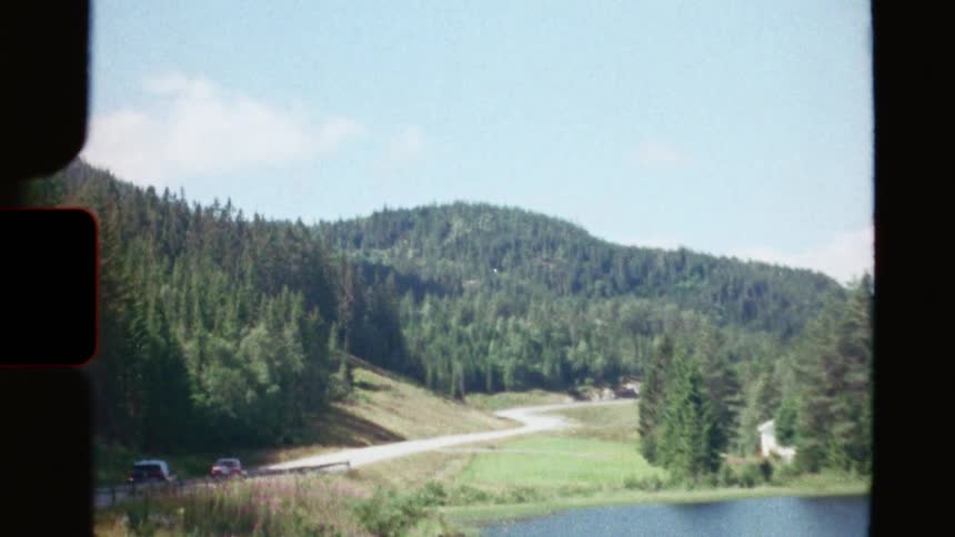Cars drive along a scenic winding road next to a tranquil lake and dense forest in this vintage Super 8 film
