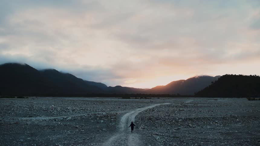 A lone adventurer runs in slow motion through a vast mountain valley towards the setting sun