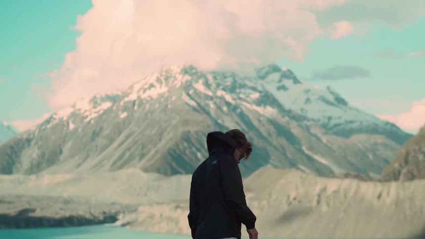 A lone adventurer walks in slow motion before a breathtaking landscape of snowy mountains and a serene glacial lake