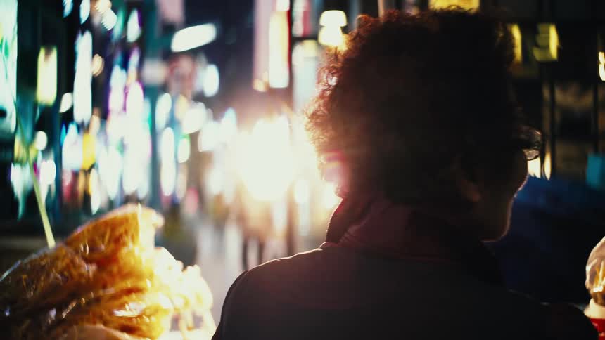 From behind, a curly-haired woman stands mesmerized by the vibrant, blurred neon lights of a bustling city street at night