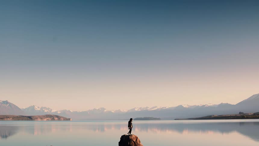 A lone photographer climbs a rock in a tranquil lake to set up a camera for a shot of the snow-capped mountains