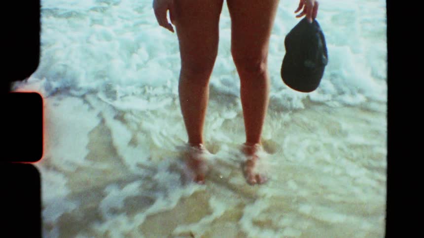 Retro film captures a woman in a bikini standing in the shallow ocean water holding a hat