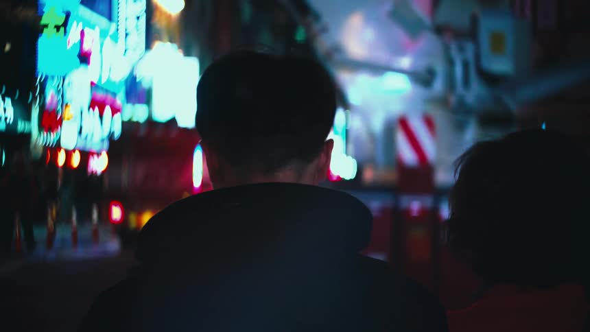 A person's silhouette is seen from behind as they walk through a bustling city street at night, with a fire truck and vibrant neon lights in the background