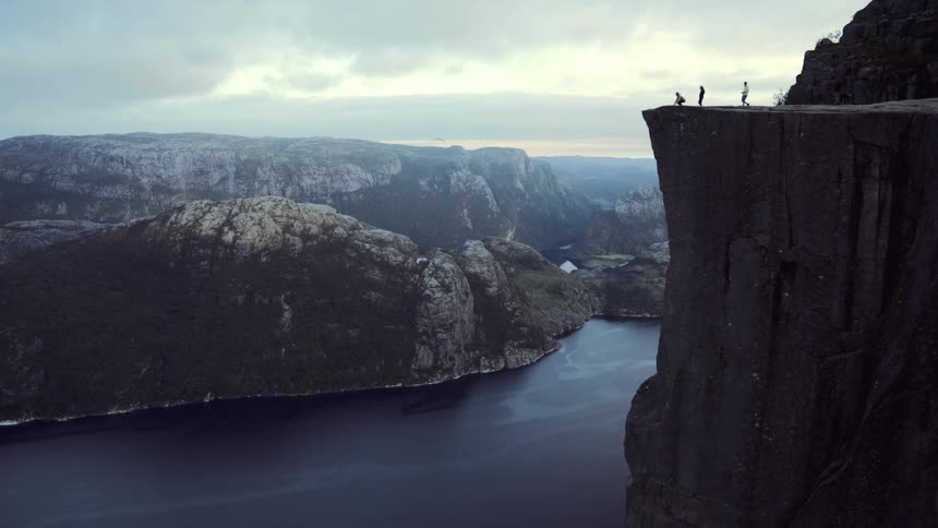 Hikers and a dog walk to the edge of the massive Preikestolen cliff, overlooking the majestic Lysefjord in Norway on a cloudy day