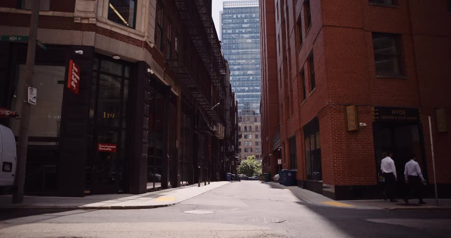 Two businessmen in white shirts and dark pants walk across an empty city street between brick buildings