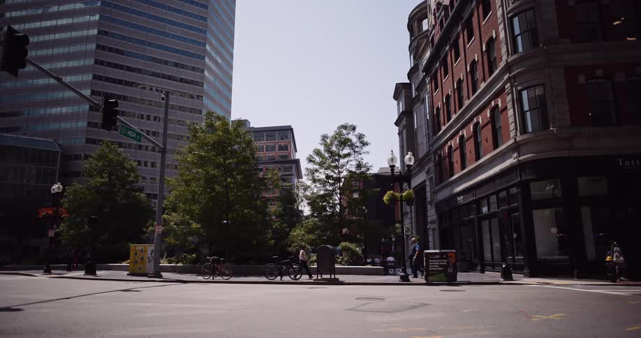 A wide static shot of a city street corner where pedestrians walk past a mix of modern and historic architecture on a bright sunny day