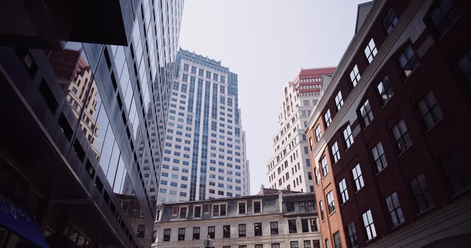 A low-angle view shows modern skyscrapers towering over older brick buildings in a downtown financial district